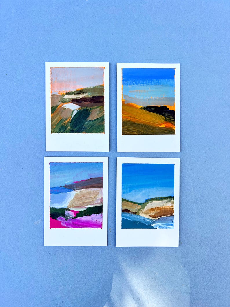 May include: Four small landscape paintings on white frames. Each painting features a different abstract landscape scene with brushstrokes of blue, green, brown, pink, and yellow. The paintings are arranged on a light blue surface.