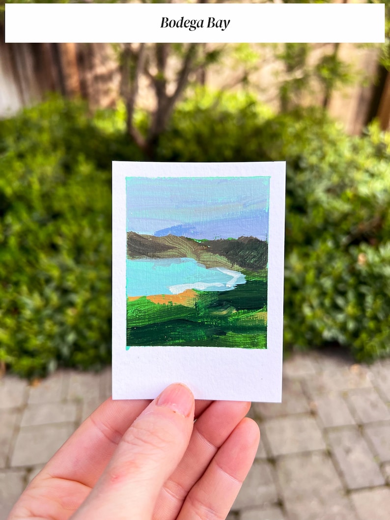 May include: A small landscape painting of Bodega Bay, featuring a turquoise bay, green hills, and a blue sky. The painting is on a white rectangular card, held by a hand, with the text "Bodega Bay" at the top.