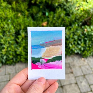 May include: A small, square painting of Montara Beach, held in a hand. The artwork features a colorful abstract landscape with blue sky, pink, green, and beige brushstrokes. The painting is on white paper, with the title "Montara Beach" above.