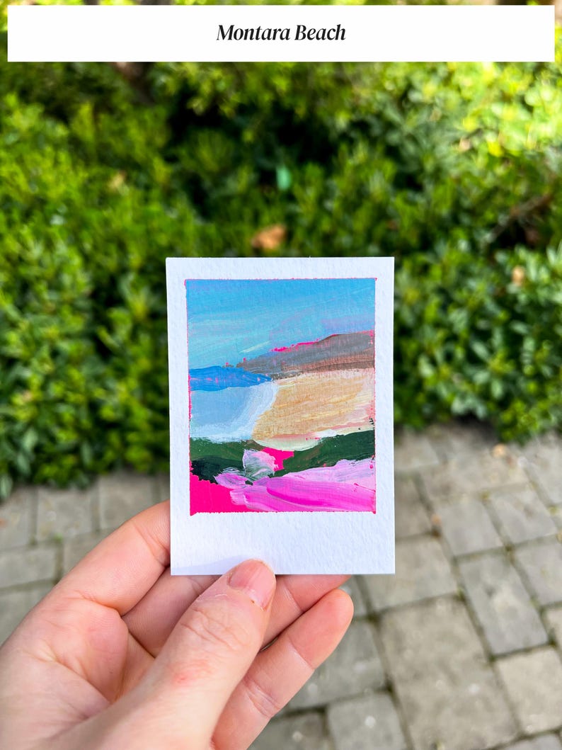 May include: A small, square painting of Montara Beach, held in a hand. The artwork features a colorful abstract landscape with blue sky, pink, green, and beige brushstrokes. The painting is on white paper, with the title "Montara Beach" above.