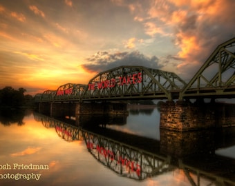 May include: A scenic photograph of a bridge over a calm river at sunset. The bridge is green with red text that reads "THE WORLD TAKES". The sky is filled with orange and yellow hues, reflecting in the water.
