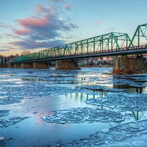 Puede incluir: Un puente de metal verde cruza un río con una superficie congelada. El río está cubierto de placas de hielo que reflejan el cielo azul y las nubes rosadas. El puente tiene una base de piedra y está rodeado de árboles.