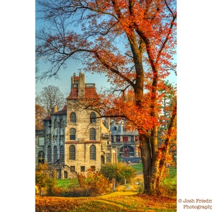 Puede incluir: Un edificio de piedra con un techo de tejas rojas y un gran árbol con hojas naranjas en primer plano. El edificio tiene muchas ventanas y una torre alta. El árbol está en otoño y las hojas están cambiando de color.