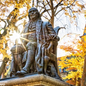 Könnte beinhalten: Eine Bronzestatue eines Mannes, der auf einem Stuhl sitzt, mit Herbstlaub im Hintergrund. Die Statue befindet sich in einem Park oder im Freien. Der Mann trägt einen langen Mantel und hat einen ernsten Gesichtsausdruck.