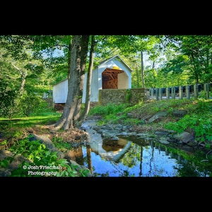 Puede incluir: Fotografía de un puente cubierto blanco con interior de madera, rodeado de árboles verdes. Un arroyo fluye en primer plano, reflejando el puente y el cielo. La imagen está enmarcada por árboles y follaje.