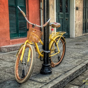May include: A yellow bicycle with a brown wicker basket and a red seat is parked in front of a building. The bicycle has a black metal frame and a black metal handlebar. The bicycle has a black metal chain and a black metal kickstand.