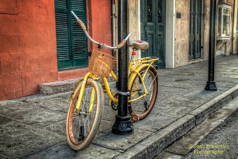May include: A yellow bicycle with a brown wicker basket and a red seat is parked in front of a building. The bicycle has a black metal frame and a black metal handlebar. The bicycle has a black metal chain and a black metal kickstand.
