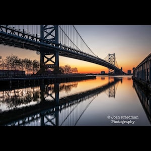 Peut inclure: Photographie d'un pont suspendu au lever du soleil. La structure en acier du pont se reflète dans l'eau calme en dessous, avec un ciel orange et jaune vibrant en arrière-plan. L'image est encadrée par des arbres et un bâtiment.