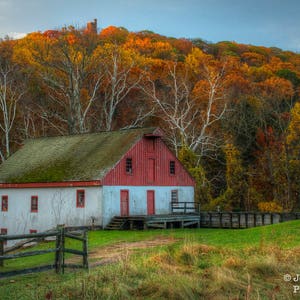 May include: A red barn with white trim sits in a field surrounded by colorful autumn trees. The barn has a gabled roof and a large door in the center. The trees are mostly shades of orange, yellow, and red.