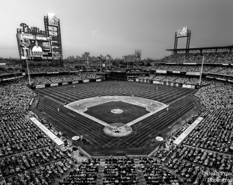 May include: A black and white photo of a baseball stadium with a large crowd of people in the stands. The field is empty and the scoreboard displays the word "Phillies".