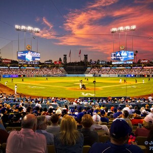 Pode incluir: Um jogo de beisebol acontecendo à noite com uma multidão de pessoas nas arquibancadas. O céu está vibrante em laranja e rosa enquanto o sol se põe. O placar mostra o número 76.
