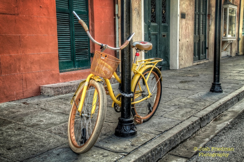May include: A yellow bicycle with a brown wicker basket and a red seat is parked in front of a building. The bicycle has a black metal frame and a black metal handlebar. The bicycle has a black metal chain and a black metal kickstand.