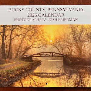 May include: A 2026 calendar featuring a scenic photograph of Bucks County, Pennsylvania. The image shows a bridge over a calm river at sunset, with trees lining the banks. The text at the top reads "Bucks County, Pennsylvania 2026 Calendar Photographs by Josh Friedman."