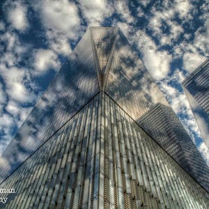 May include: A low angle view of a tall, modern skyscraper with a glass facade reflecting the cloudy sky. The building is made up of many vertical panels of glass, creating a grid-like pattern. The sky is a mix of white and gray clouds.
