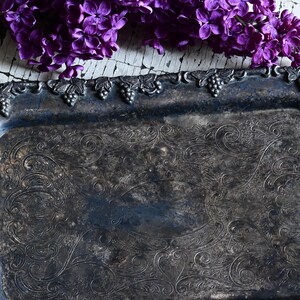 May include: An antique silver-toned rectangular tray with ornate scrollwork and grape cluster detailing along the edges. The tray is tarnished, giving it a vintage appearance. A sprig of purple lilacs rests above the tray.