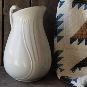 May include: A vintage white ceramic pitcher with a curved handle and decorative vertical lines. The pitcher is next to a folded quilt with blue and brown triangle patterns on a white background. The background is a weathered wooden surface.