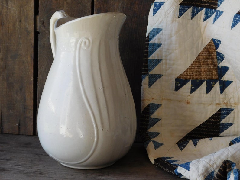 May include: A vintage white ceramic pitcher with a curved handle and decorative vertical lines. The pitcher is next to a folded quilt with blue and brown triangle patterns on a white background. The background is a weathered wooden surface.