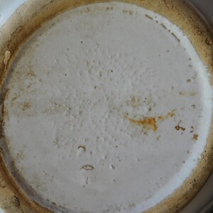 May include: Close-up of the bottom of a white ceramic bowl. The bowl's interior is a smooth, off-white color, while the rim and base have a textured, light brown appearance. The bowl shows signs of wear and age.
