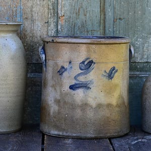 May include: Three antique stoneware crocks of varying sizes and shapes. The center crock is cylindrical with blue painted floral designs. The other two are a light beige color. The crocks are displayed on a weathered wooden surface.