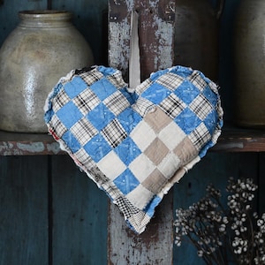 May include: A quilted heart-shaped decoration with a patchwork design in shades of blue, white, and tan. The heart is hanging against a weathered wooden background, with antique stoneware jars on a shelf behind it.