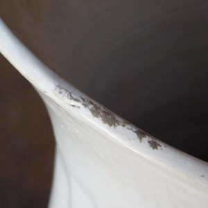 May include: Close-up of a white ceramic vase with a slightly flared rim. The rim shows signs of wear with small areas of discoloration. The interior of the vase is dark, contrasting with the bright white exterior.
