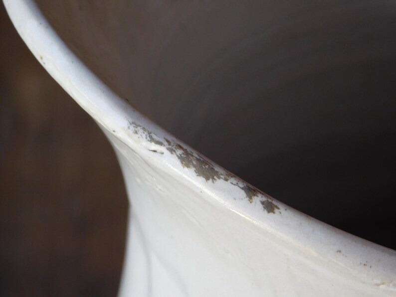 May include: Close-up of a white ceramic vase with a slightly flared rim. The rim shows signs of wear with small areas of discoloration. The interior of the vase is dark, contrasting with the bright white exterior.