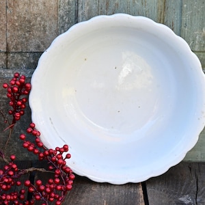 May include: A white, scalloped-edge bowl sits on a weathered wooden surface, with a backdrop of aged wooden planks. Red berry branches with small, round berries are arranged to the left of the bowl, adding a pop of color.