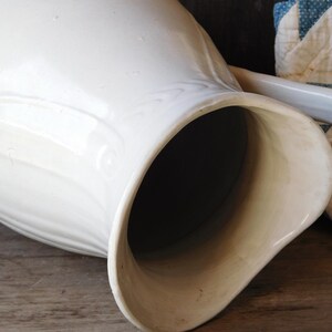 May include: A large, off-white ceramic pitcher with a curved handle and a wide spout. The pitcher has a smooth, glossy finish and a slightly textured surface. The interior is dark. The pitcher is likely vintage or antique.