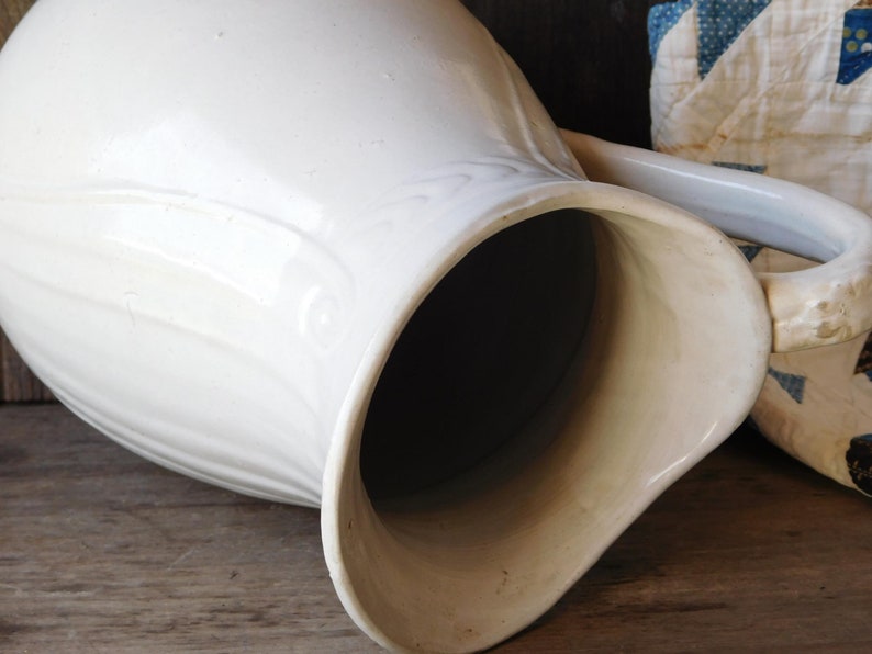 May include: A large, off-white ceramic pitcher with a curved handle and a wide spout. The pitcher has a smooth, glossy finish and a slightly textured surface. The interior is dark. The pitcher is likely vintage or antique.