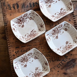 May include: Four square, off-white ceramic dishes with a brown floral pattern. The dishes are arranged on a rustic wooden surface, creating a vintage aesthetic. The floral design adds a touch of elegance to the simple, square shape.