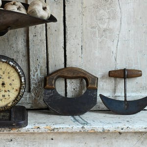 May include: A vintage dark metal kitchen scale with a round cream-colored dial and two crescent-shaped metal blades with wooden handles. The scale features a large metal pan on top and a circular face with black numbers and a central needle indicator.