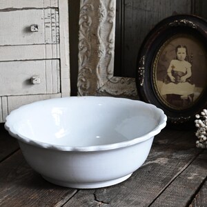 May include: A white ceramic bowl with a scalloped rim sits on a rustic wooden surface. The bowl is the focal point, reflecting light. In the background, a vintage framed portrait and a white cabinet add to the antique aesthetic.