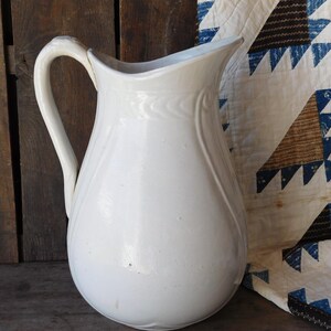 May include: A white ceramic pitcher with a curved handle and a spout. The pitcher has a rounded body and a slightly textured surface. The background includes a wooden surface and a quilt with blue and brown triangle patterns.