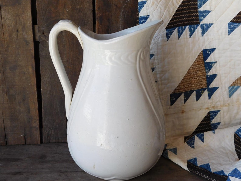 May include: A white ceramic pitcher with a curved handle and a spout. The pitcher has a rounded body and a slightly textured surface. The background includes a wooden surface and a quilt with blue and brown triangle patterns.