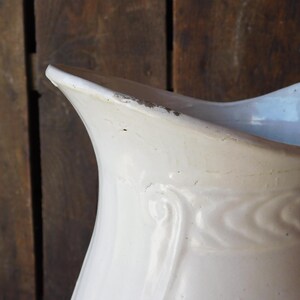 May include: Close-up of a white ceramic pitcher with a curved spout and a decorative embossed pattern. The pitcher has a slightly distressed appearance, with visible wear and tear along the edges, suggesting age and use. The background is a rustic wooden surface.