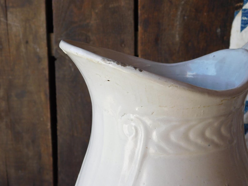 May include: Close-up of a white ceramic pitcher with a curved spout and a decorative embossed pattern. The pitcher has a slightly distressed appearance, with visible wear and tear along the edges, suggesting age and use. The background is a rustic wooden surface.