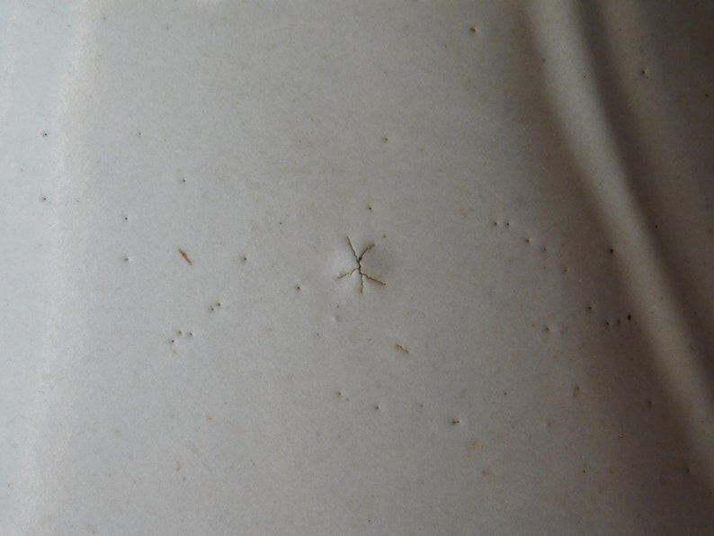 May include: Close-up of a textured, off-white surface with small, dark imperfections. A star-shaped crack is visible near the center. The surface appears to be painted or coated.