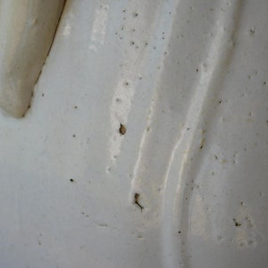 May include: Close-up of a white ceramic pitcher with a handle. The pitcher has a glossy finish with subtle texture and small dark spots. The handle is a lighter shade of white.