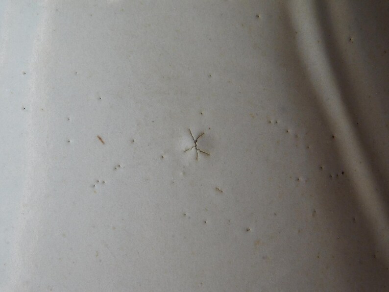 May include: Close-up of a textured, off-white surface with small, dark imperfections. A star-shaped crack is visible near the center. The surface appears to be painted or coated.
