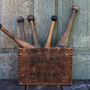 May include: A wooden box filled with antique wooden exercise clubs. The clubs have rounded heads and tapered handles. The box has text that reads "Hardy Box French Pine Drinks". The background is a weathered, teal-colored wooden door.
