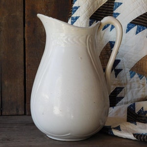 May include: A white ceramic pitcher with a curved handle and a spout. The pitcher has a bulbous body and decorative detailing near the top. The background includes a wooden surface and a patterned quilt.