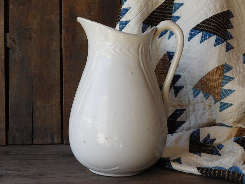 May include: A white ceramic pitcher with a curved handle and a spout. The pitcher has a bulbous body and decorative detailing near the top. The background includes a wooden surface and a patterned quilt.
