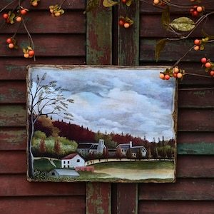 May include: A framed painting depicting a rural landscape with a small village nestled among trees. The sky is a soft blue with fluffy white clouds. The trees are a mix of green and brown, and the houses are white with red roofs. The painting is set against a rustic wooden background.