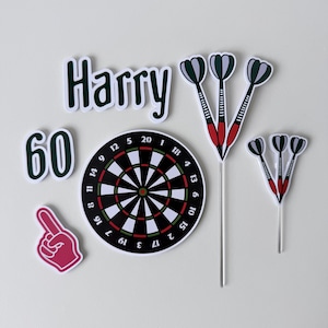 May include: A set of cake toppers featuring the name Harry in dark green, the number 60, a circular dartboard, a pink foam finger, and two clusters of three darts on sticks. The dartboard displays standard black, white, and red scoring segments.