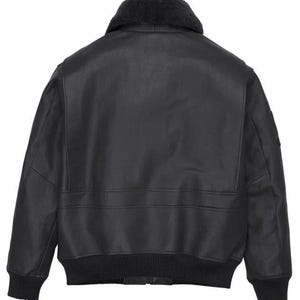 May include: A black leather bomber jacket featuring a dark gray faux fur collar, long sleeves with ribbed cuffs, and a ribbed hem. The back of the jacket displays a smooth leather finish with horizontal seam detailing across the mid-section.