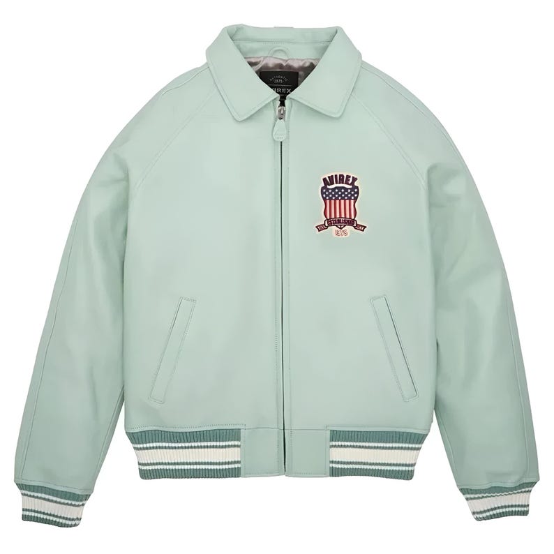 May include: A light mint green jacket with a full-length front zipper, a classic collar, and ribbed cuffs and hem featuring white stripes. The chest displays an embroidered patch with the text AVIREX and a shield graphic with a flag design.