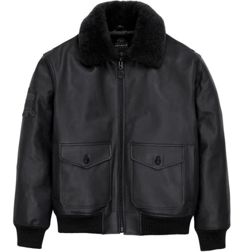 May include: A black leather bomber jacket featuring a black faux fur collar, a central zipper closure, and two front flap pockets with button fasteners. The jacket includes ribbed cuffs and hem, with a small patch detail on the right sleeve.