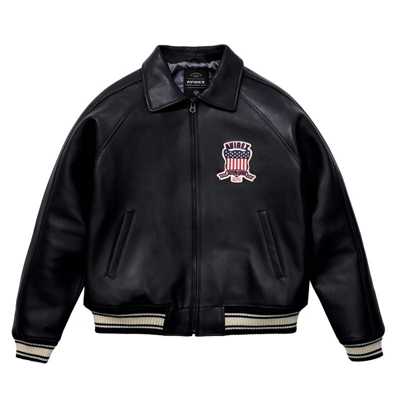 May include: A black leather jacket with a classic collar, a full-length front zipper, and ribbed cuffs and hem featuring white stripes. On the left chest, there is a patch displaying the text AVIREX above a shield with a red, white, and blue flag design.