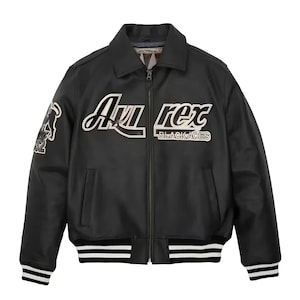 May include: A black leather bomber jacket featuring a front zipper closure, a collar, and striped ribbed cuffs and hem. The chest displays white and black embroidered text reading Avirex Black Aces, with a graphic patch on the right sleeve.