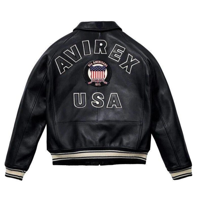 May include: A black leather varsity jacket featuring the white embroidered text AVIREX and USA on the back. A circular patch in the center displays the American flag with the text ALL AMERICANS ESTABLISHED 1975. The jacket has striped ribbed cuffs.
